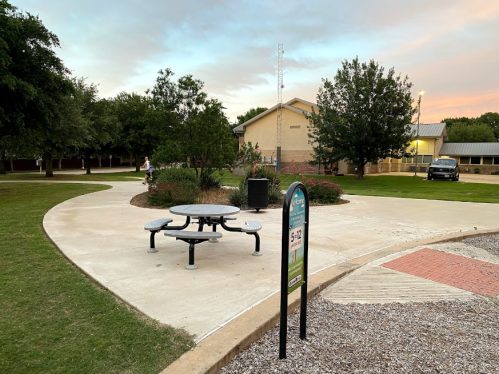 A park scene featuring a picnic table, grassy area, and a building in the background under a colorful sky.