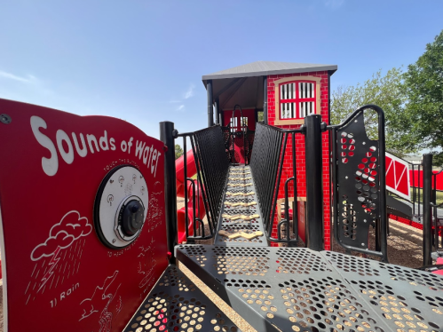 A colorful playground structure with a "Sounds of Water" panel, featuring a bridge leading to a playhouse.