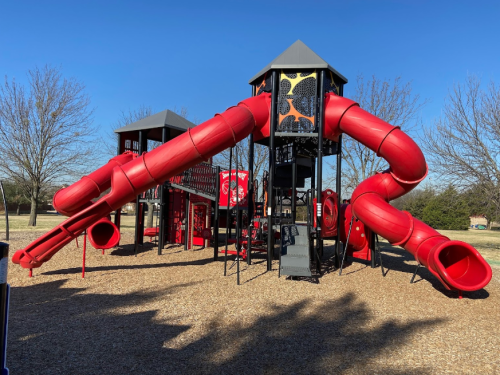 A colorful playground featuring large red slides and a play structure, set in a grassy area with trees in the background.