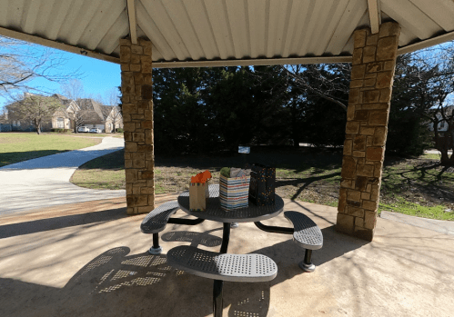 A shaded picnic area with a table, surrounded by trees and a clear blue sky, featuring colorful gift bags on the table.