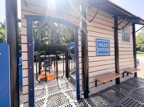 A playground structure resembling a police station with a sign, benches, and climbing equipment in the background.