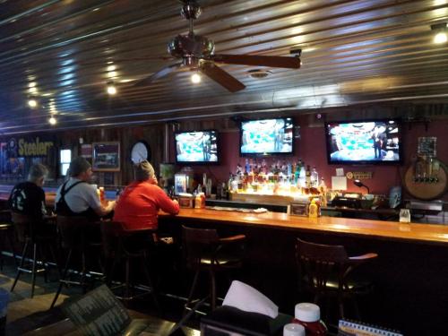 A cozy bar with patrons seated at the counter, two TVs showing sports, and a well-stocked shelf of liquor behind the bar.