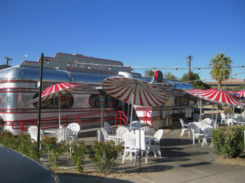 A retro diner with a silver exterior, red and white striped umbrellas, and outdoor seating under a clear blue sky.