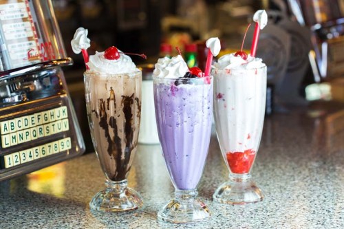 Three colorful milkshakes in tall glasses, topped with whipped cream and cherries, on a speckled countertop.