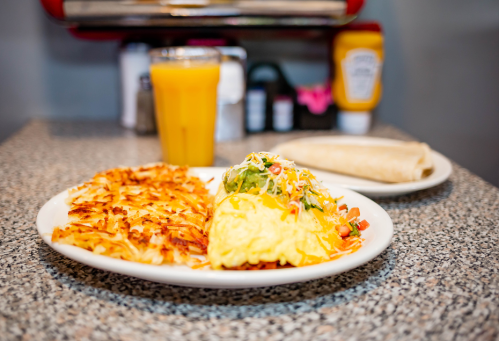 A plate of fluffy omelette with toppings, crispy hash browns, and a glass of orange juice on a speckled table.
