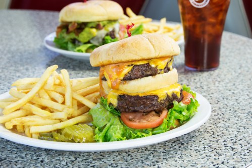 A stacked cheeseburger with lettuce, tomato, and fries on a plate, with a drink in the background.