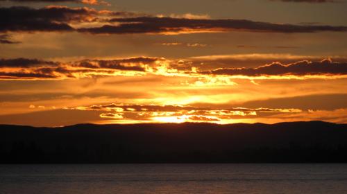 A vibrant sunset over a calm lake, with golden and orange hues reflecting on the water and silhouetted mountains in the background.
