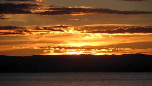 A vibrant sunset over a calm lake, with golden and orange hues reflecting on the water and silhouetted mountains in the background.