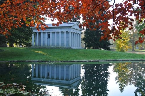 A classical building with columns reflected in a calm pond, surrounded by vibrant autumn foliage.