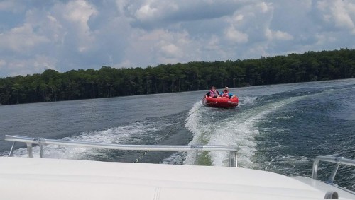 Two people on a red inflatable tube being towed on a lake, with trees and clouds in the background.