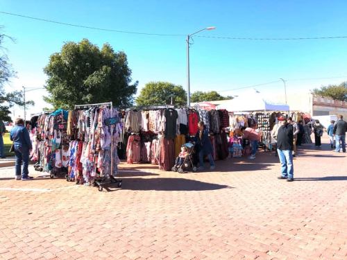 A bustling outdoor market with clothing racks and shoppers browsing among colorful garments on a sunny day.
