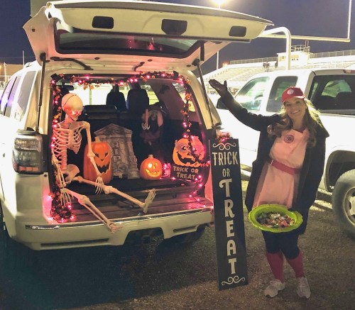 A woman in a pink outfit stands by a decorated SUV trunk with Halloween decorations, including a skeleton and pumpkins.