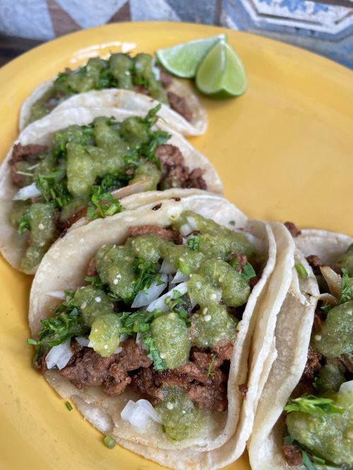 A plate of tacos filled with beef, topped with green salsa, onions, and cilantro, served with a lime wedge.