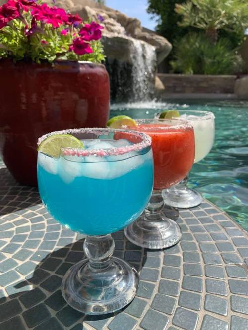 Three colorful cocktails with lime slices and salt rims sit on a table by a pool, with flowers and a waterfall in the background.