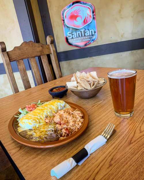 A plate of enchiladas with rice, a bowl of salsa, tortilla chips, and a pint of beer on a wooden table.