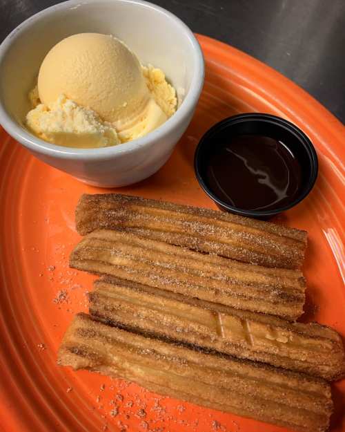 A plate with three churros dusted in sugar, a scoop of vanilla ice cream, and a small cup of chocolate sauce.