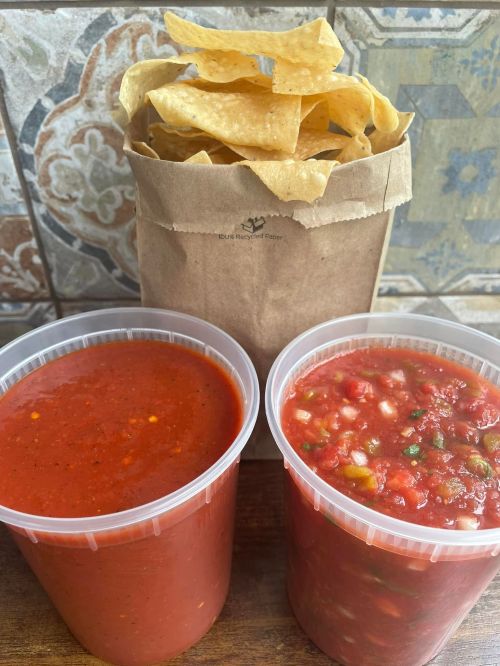 Two containers of salsa, one red and one chunky, beside a bag of tortilla chips. Background features decorative tiles.