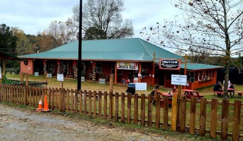 A rustic restaurant with a green roof, outdoor seating, and a wooden fence, surrounded by trees and a gravel path.