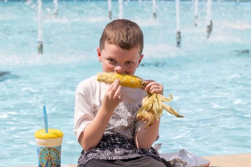 A boy sitting by a pool, eating corn on the cob, with a drink beside him and a playful expression.