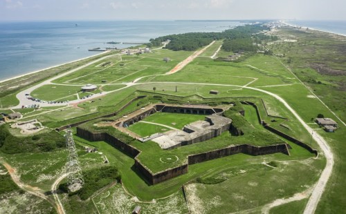 Aerial view of a historic fort surrounded by green fields and a coastline, with a road and parking area nearby.