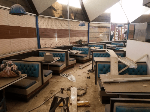 Abandoned restaurant interior with overturned furniture, debris, and dusty booths under dim lighting.