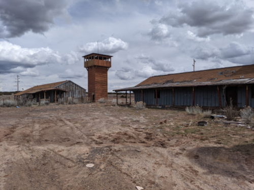 Abandoned buildings with a watchtower under a cloudy sky, surrounded by barren land.