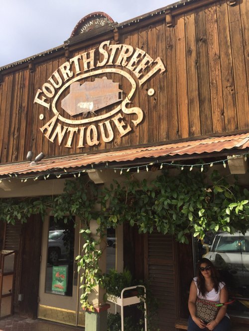 A woman sits outside a rustic antique shop with a wooden sign reading "Fourth Street Antiques" surrounded by greenery.
