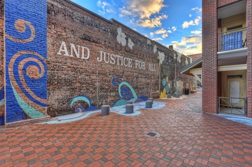 Mural on a brick wall featuring the phrase "AND JUSTICE FOR ALL" with colorful swirls and a paved walkway.