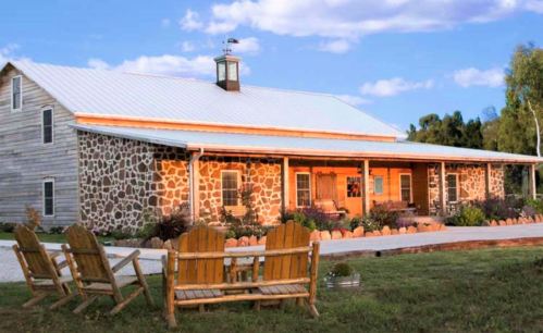 A rustic stone and wood house with a metal roof, surrounded by greenery and featuring wooden chairs in front.