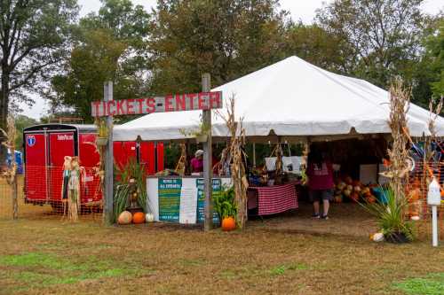 A tent labeled "TICKETS ENTER" with pumpkins and decorations, set in a grassy area with trees in the background.