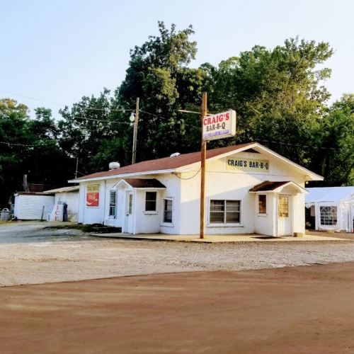 A small, white building with a sign reading "Craig's Bar-B-Q," surrounded by trees and gravel.