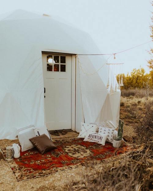 A cozy white dome tent with a door, surrounded by pillows and a rug, set in a natural outdoor landscape.