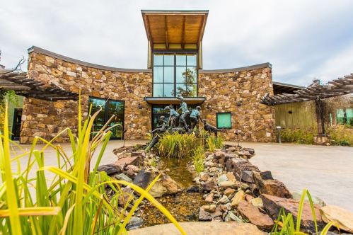 A modern stone building with large windows, featuring a sculpture and a small water feature in the foreground.