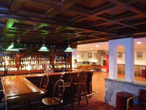 A cozy bar with wooden decor, green pendant lights, and a stocked liquor shelf, featuring wooden chairs and a warm atmosphere.