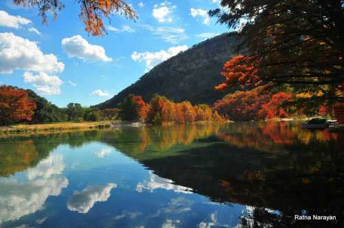 A serene lake reflects colorful autumn trees and a blue sky with fluffy clouds, framed by a mountain backdrop.