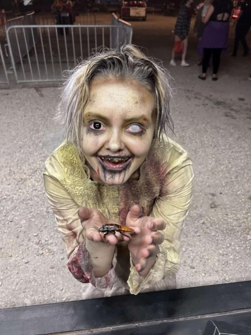 A zombie-like figure with messy hair and eerie makeup holds out her hands, smiling in a spooky setting.