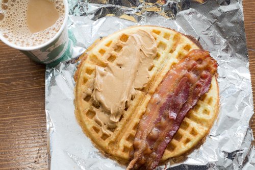 A waffle topped with peanut butter and crispy bacon, alongside a cup of coffee on a wooden table.