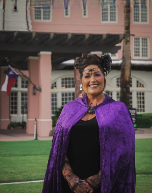 A woman in a purple cape smiles outdoors, with a historic building and Texas flag in the background.