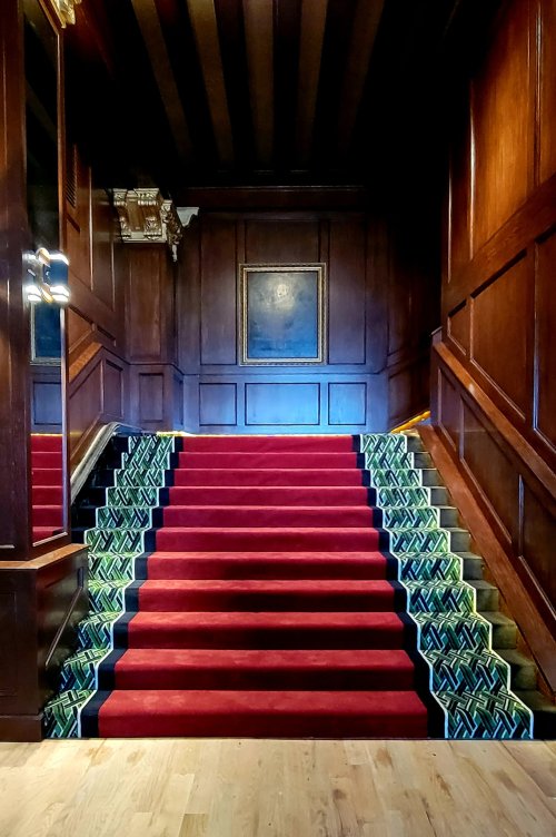 A grand staircase with red carpet and patterned edges, surrounded by dark wooden walls and a framed painting above.