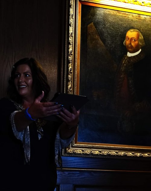 A woman gestures while standing next to a large portrait of a man in historical attire, illuminated by soft light.