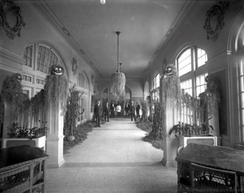 A historic hallway decorated with plants and figures, featuring large windows and people in formal attire.