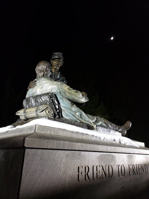A bronze statue depicting two soldiers embracing, with a moonlit sky in the background. Inscription reads "FRIEND TO FRIEND."