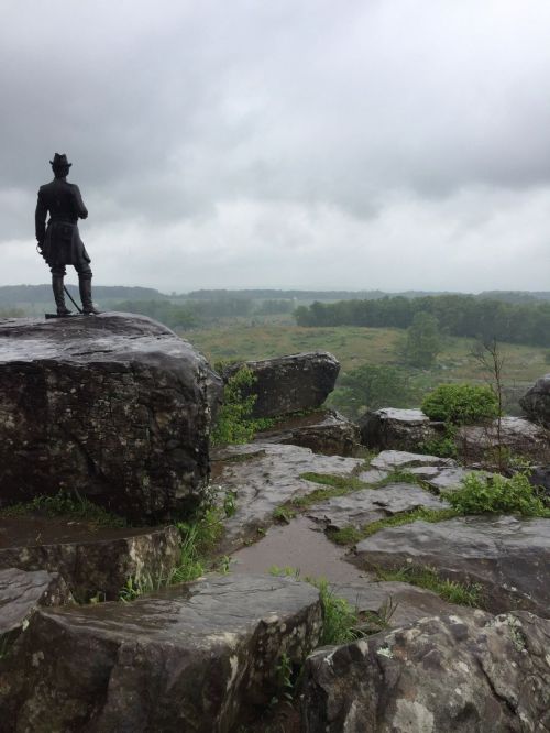 A statue stands on a rocky outcrop overlooking a misty, green landscape under a cloudy sky.