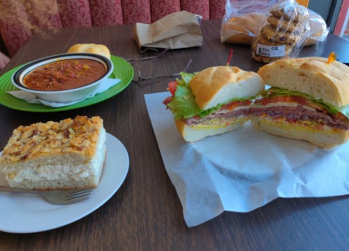 A table with a sandwich, a bowl of chili, a slice of cake, and rolls in the background.