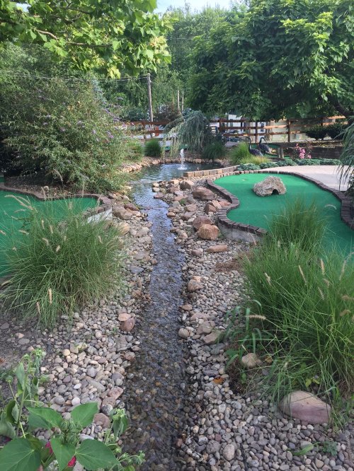 A serene landscape featuring a winding stream, rocks, and lush greenery, with a mini-golf course in the background.