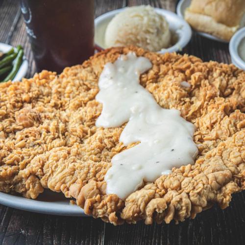 A large, crispy fried chicken breast topped with creamy white gravy, served with sides of mashed potatoes and green beans.