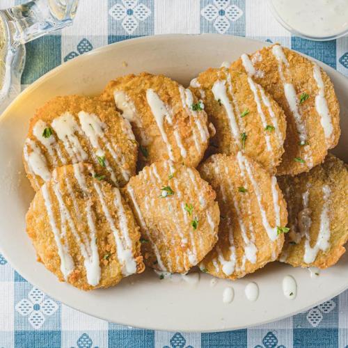 A platter of crispy fried green tomatoes drizzled with creamy sauce, garnished with herbs, on a checkered tablecloth.