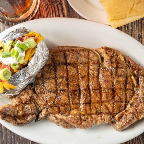 A grilled steak with grill marks, served with a baked potato topped with cheese, bacon, and green onions, and a roll.