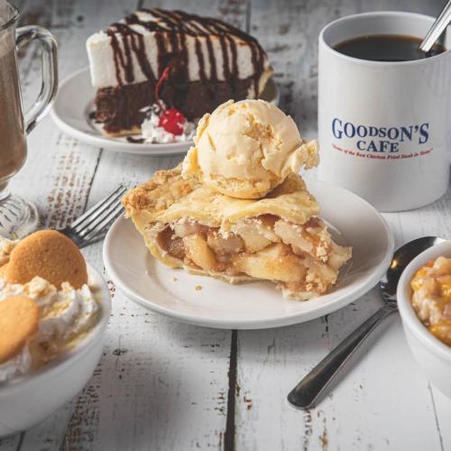 A slice of apple pie topped with ice cream, surrounded by coffee, chocolate cake, and dessert bowls on a rustic table.