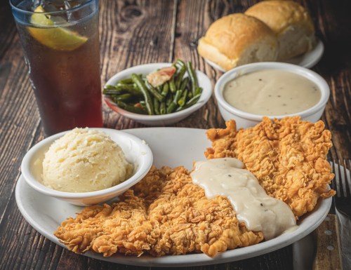 A plate of fried chicken with gravy, mashed potatoes, green beans, and rolls, alongside a glass of iced tea.